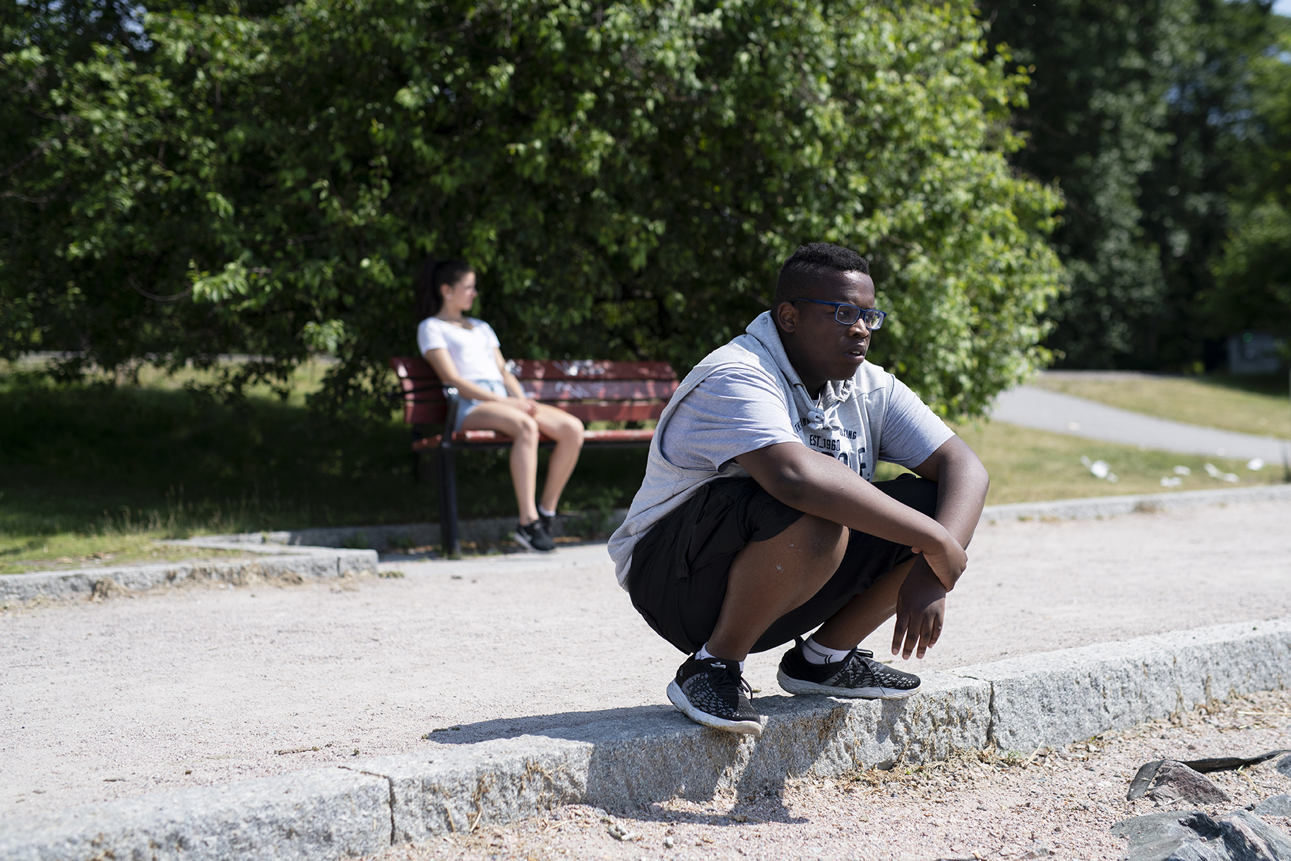 Nuori mies istuu kyykyssä jalkakäytävän reunalla, taustalla hahmo penkillä.