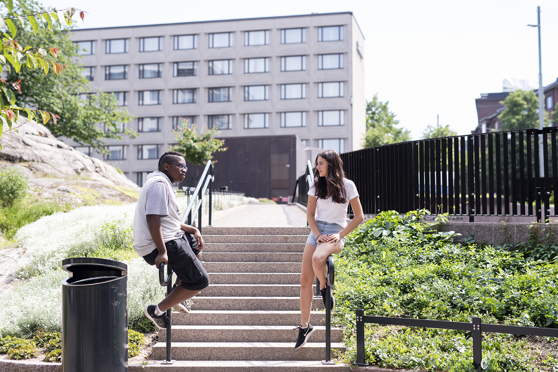 Poika ja tyttö istuvat ulkoportaikon aidalla kesäisessä kaupungissa.