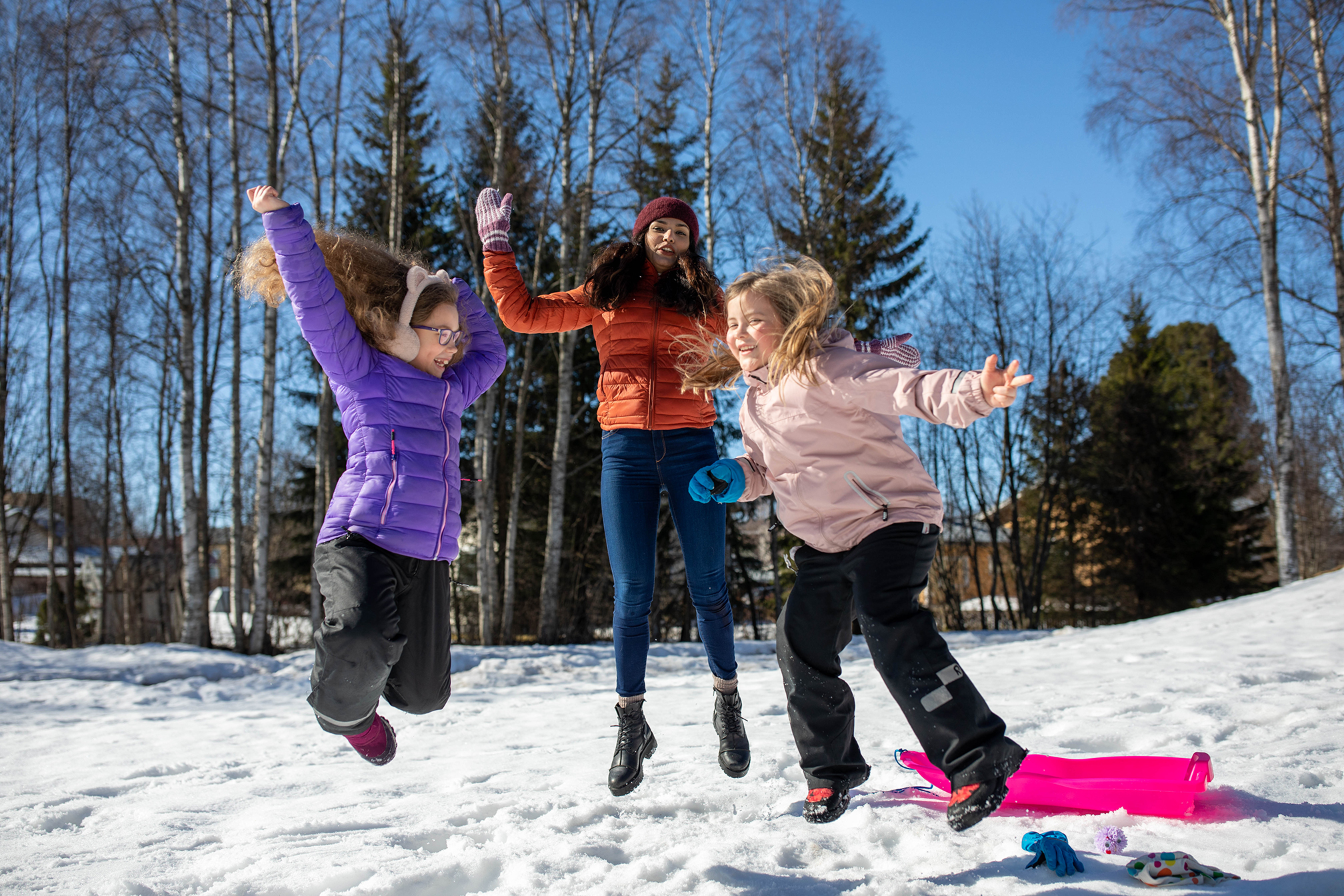 Kaksi lasta ja aikuinen värikkäissä toppatakeissa hyppää ilmaan. Maassa on lunta ja taustalla metsää ja sininen taivas.