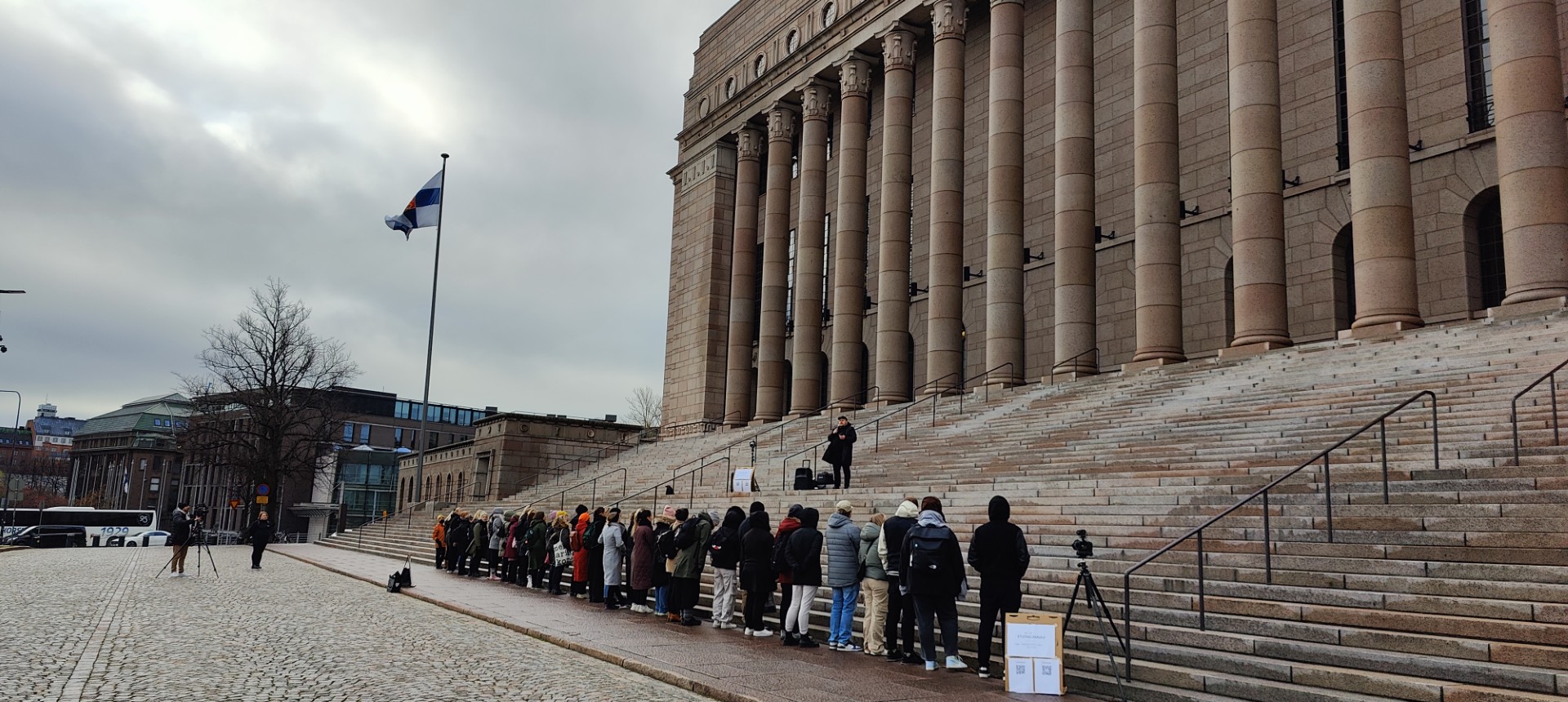 Ihmisiä rivissä Eduskuntatalon portaiden alapäässä.