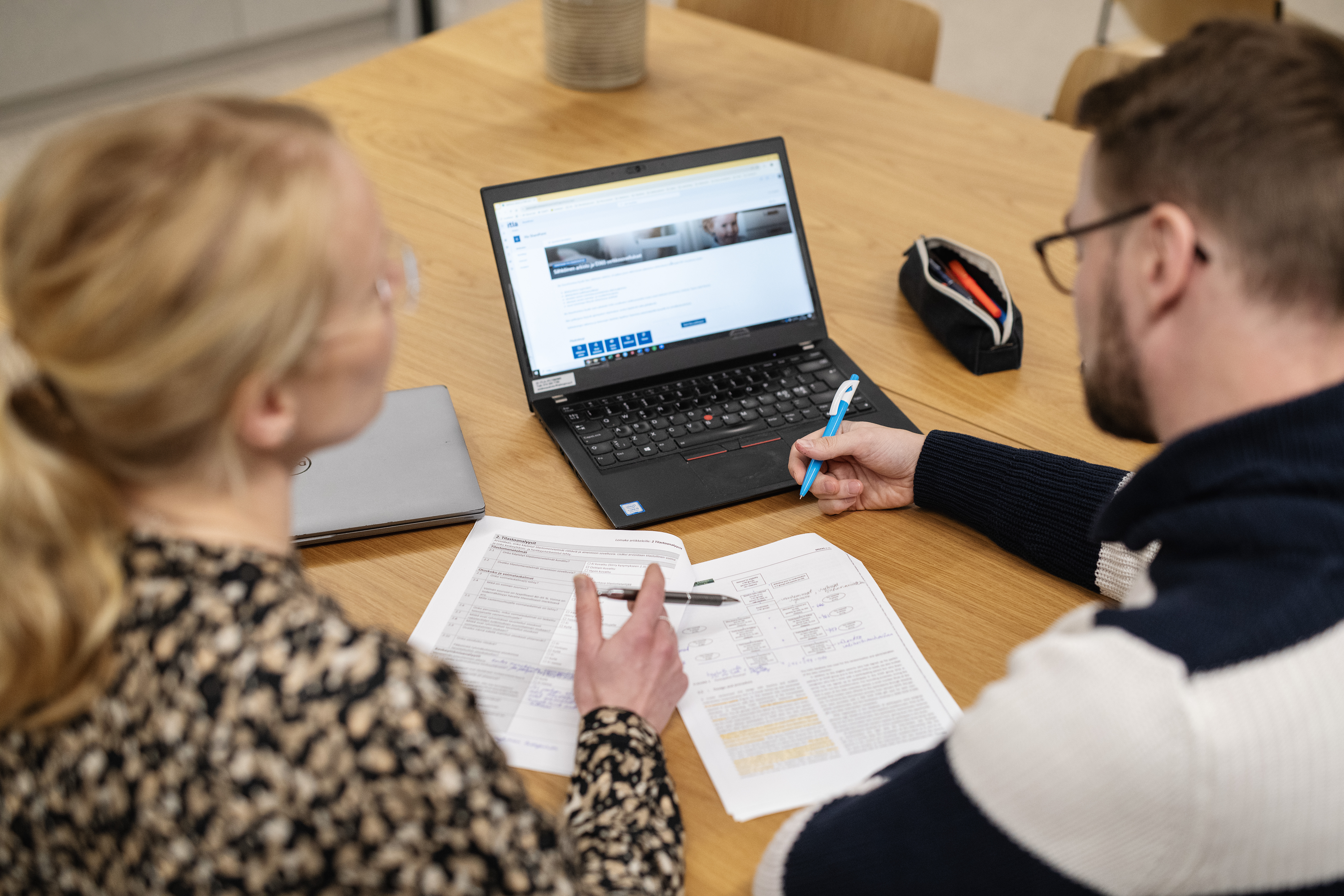 Kaksi ihmistä (toisella pitkät vaaleat hiukset ja toisella tummat hiukset ja parta) keskustelevat läppärin äärellä.