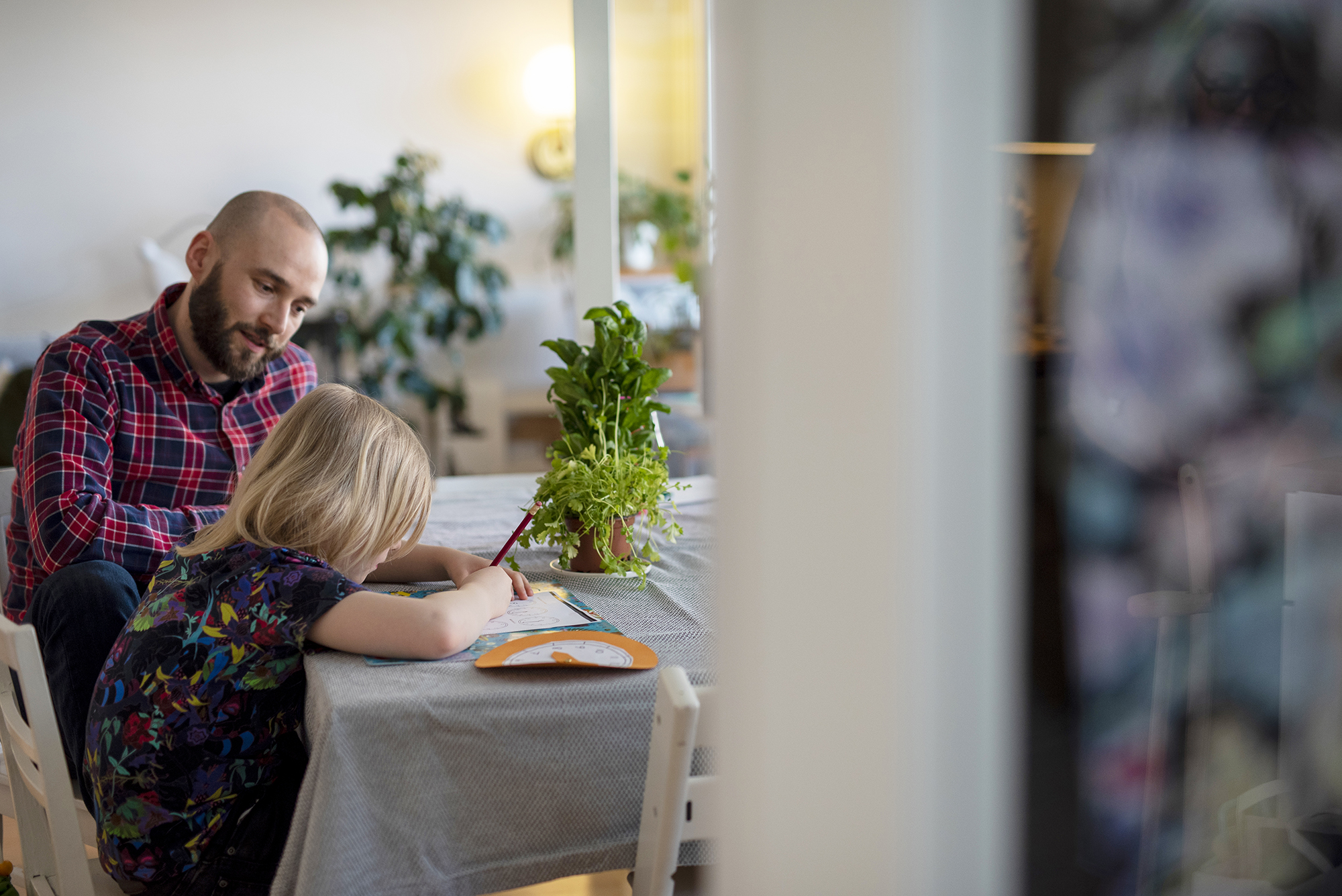 Ruutupaitainen isä ja sinipaitainen lapsi istuvat keittiön pöydän ääressä. Lapsi piirtää.