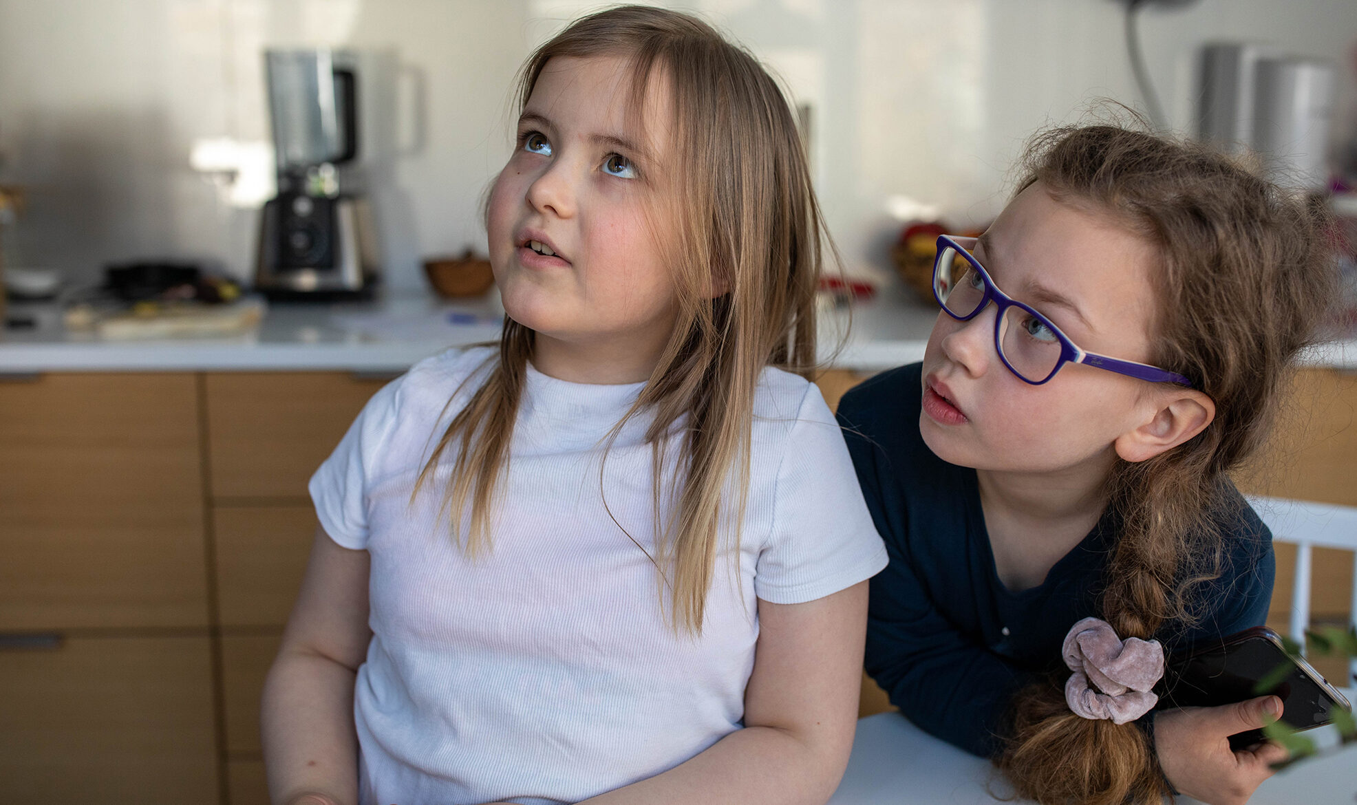 Kaksi lasta katsoo ylöspäin. Toisella vakoinen paita ja pitkät hiukset auki, toisella tumma paita, letti ja silmälasit