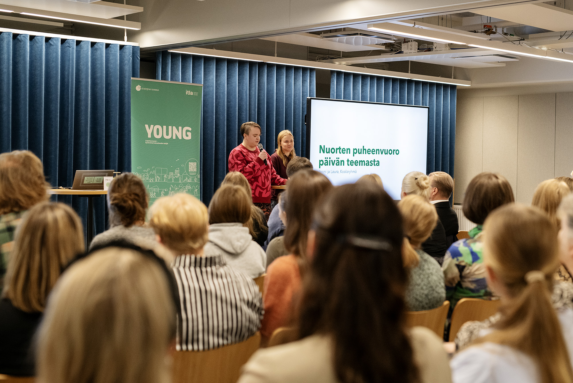 Yleisö kuuntelee kahden nuoren puheenvuoroa seminaarissa.